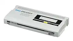 Photographie d'une console rectangulaire blanche, avec une partie noire sur le côté supérieur droit, et où est inscrit Sega en bleu à gauche.