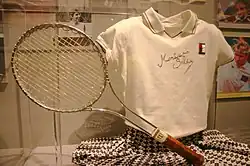 Photo de la tenue de Seles à son retour en 1995. Polo blanc dédicacé, jupe à carreaux bleu marine et blanc.