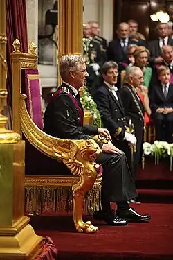 Photographie de profil d'un homme aux cheveux blancs assis sur un fauteuil aux accoudoirs dorés