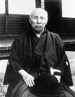 photo d'un homme chauve habillé en costume traditionnel japonais, assis en tailleur.