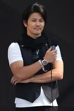 Photographie d'un commentateur sportif engilet noir, tenant un micro.