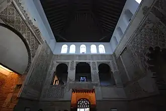 La photo panoramique, prise en contre-plongée, montre trois des quatre murs de la salle de la synagogue, avec au centre le mur sud, ainsi que son plafond fait d'une voûte de bois sombre. Les murs, d'une couleur blanc cassé, sont ornés de bas-reliefs et d'arches de style mudéjar et d'inscriptions en hébreu.