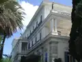 Vue de l'Achilleion laissant apparaître l'une des muses du premier étage ainsi que l'Hermès ailé (façade gauche du bâtiment principal)