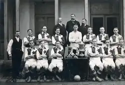 Photographie en noir et blanc d'une équipe de football avec treize joueurs, trois dirigeants, et l'entraîneur à gauche, tous de face.