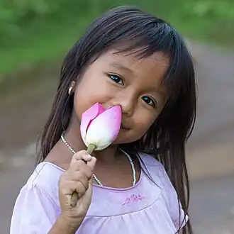 Fillette laotienne souriante fixant l'observateur, la tête inclinée vers la gauche et tenant un bourgeon de fleur de lotus dans sa main droite.