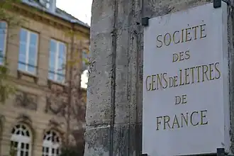 Photographie en couleur d'une plaque sur un pan de mur, blanche avec des caractères dorés