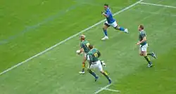 Vue de loin et de derrière d'un joueur en vert s'apprêtant à marquer un essai, suivi par deux coéquipiers, un adversaire en bleu tentant de couper sa course par le côté.