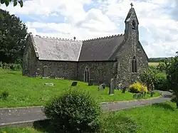 Photo d'un bâtiment en L avec un petit clocher. Une allée goudronnée y conduit en serpentant au milieu d'une pelouse jonchée de pierres tombales.