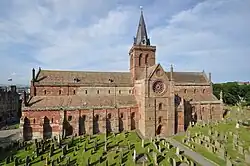 Long édifice religieux en pierres, avec entrée par le transept, entouré de nombreuses stèles funéraires.