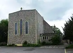 Photo d'un bâtiment à la façade de pierre grise imposante, percée de trois fenêtres en ogive, surplombé d'une croix chrétienne