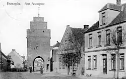 Photo en noir et blanc montrant une rue du début du XXe siècle. À l'arrière plan, on voit une tour percée d'une grande porte.