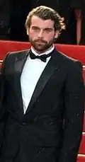 Photo de faible résolution montrant un homme debout sur des escaliers rouges, vêtu d'un costume noir, d'une chemise blanche et d'un nœud papillon.