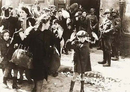 Dans une rue, des civils (des femmes et des enfants au premier plan, quelques hommes derrières) ont les mains levés. Derrière eux, des soldats ont leurs armes pointés sur les civils et font sortir d’autres personnes d’un bâtiment.