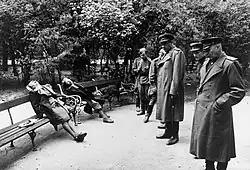Six hommes en uniforme regardent trois cadavres reposant sur les bancs d'un jardin public. L'un de ces corps est couché, tandis que les deux autres sont assis, la tête basculée en arrière ou sur le côté.