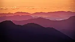 Une série de crêtes de montagnes, de noir, violet, un ciel orange, quelques rayons de soleil rasent les vallées.