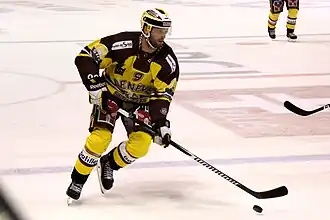 Hockeyeur en pied, de face, en train de patiner, la crosse posée sur la glace sur sa gauche