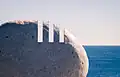 Photo agrandie du coin supérieur droit de l'une des pierres commémoratives. Trois entailles droites y sont incrustées, représentant le chiffre 111. À l'arrière-plan, la mer baigne au pied d'un ciel blanc de soirée.