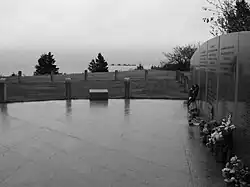 Photo présentant une vue d'un mémorial au bord de la côte. Un sol en pierre précède une vue sur le large et l'océan. À droite, un mur comporte les noms des victimes. Des fleurs sont présentes au pied du mur.