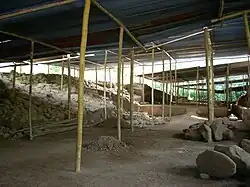 Un espace vierge comportant de la terre nue et des décombres, avec des espaces surélevés à gauche et à droite, le tout sous un toit en tôle avec une charpente en bois.