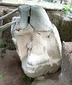 Une sculpture en pierre reposant dans une tranchée creusée. Le monument est endommagé avec une fracture profonde vers le haut. Il penche vers la gauche et est soutenu par des poteaux en bois. Une niche est creusée sur le devant du monument, et contient une silhouette assise, très érodée, avec quelque chose dans ses bras.