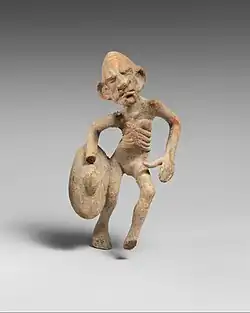 Petite statuette d'un homme vu de face, visage masqué grimaçant, corps légèrement penché vers sa droite, une jambe levée pliée devant lui, tenant un bouclier au bout du bras droit plié.