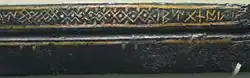 Un gros plan sur le seax montrant plus clairement une série de chevrons et de runes de couleur dorée au-dessus du sillon central.