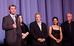 Photographie de trois hommes et deux femmes debout devant un écran de projection. L'homme à gauche sourit en parlant dans un micro.