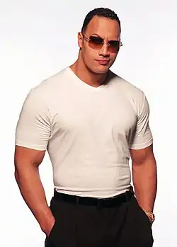 Un catcheur musclé au t-shirt blanc et au pantalon noir avec des lunettes de soleil pose devant un fond blanc avec ses mains dans les poches.