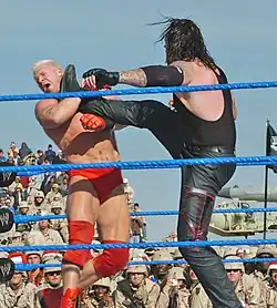 Un catcheur brun habillé en noir donne un coup de pied au visage à son adversaire, un blond en slip rouge.