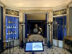 Boudoir, vers 1925, appartement privé de l'hôtel particulier de Jeanne Lanvin à Paris (démoli en 1965). Murs recouverts de lambris moulurés, surmontés de bas-reliefs en stuc; alcôve encadrée de colonnes engagées en marbre veiné avec base de bois sculpté; plinthe en bois sculpté; sol en dalles de marbres blanc et noir; vitrines d'angles tapissées de soie "bleu Lanvin". Canapé, vers 1925, avec encadrement en chêne mouluré, dossier et accotoirs enveloppants, rembourrés, capitonnés et recouverts d'une garniture d'origine de soie jaune tilleul; paire de luminaires lampadaire, vers 1924, bronze fondu et patiné vert antique, provenant de la salle de bains de Jeanne Lanvin.