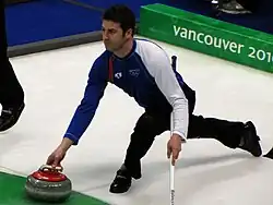 un curleur en pantalon noir, sweater bicolore blanc et bleu s’apprête à lâcher sa pierre de la main droite : large fente des jambes, bras gauche en appui sur son balai, en arrière plan marquage Vancouver 2010 en blanc sur vert