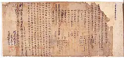 Morceau de papier bruni et déchiré aux bordures recouvert de caractères chinois à l’encre noire.