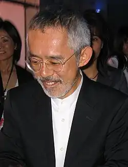 Buste et tête d'un homme asiatique d'environ 50 ans. Cheveux et barbe grisonnants, lunettes rondes.