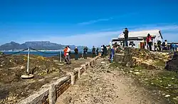 A straight stone pathway through a rocky area, elevated in places from the walkway, with a low retaining wall and chain fence on the left. Beyond is an area of ocean; at the far background on the left is a large flat rocky mountain with a peaked one at its left. Closer to the camera is a small building on the right; people are milling around it and the paths, some taking pictures