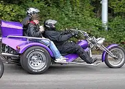 Photo couleur d'un tricycle motorisé, avec un conducteur casqué et deux passagers.