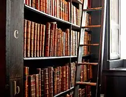 De nombreux livres anciens dans une haute étagère de bois sombre, avec une grande échelle de bois sur la droite permettant d’attraper les livres en hauteur
