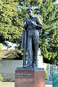 Photo couleur d'une statue d'homme en pied, regardant ses mains, devant un grand arbre