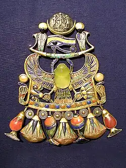 bijou avec un scarabé en verre transparent, enchassé dans du métal émaillé: connecté au scarabé sont deux ailes d'aigle déployées sur lesquelles vogue une barque stylisée surmontée du hyéroglyphe de l'oeil, et d'un croissant lunaire dans lequel est placé un sceau rond avec 3 personnages, celui du centre sous les bras levés des deux autres. Sous le sacarabé, deux pattes d'aigle tiennent à droite un bouquet de plantes - peut être du papyrus et à gauche une fleur. des deux cotés des pattes, deux cobras stylisés et en dessous une rangée alternée de pendants de formes diverses régulièrement agencés.