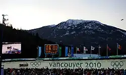 Une foule multicolore regarde passer un bobsleigh noir et blanc sur une piste de bobsleigh marquée des anneaux olympique et de grandes lettres « vancouver 2010 » ; derrière, se trouve un écran géant vidéo retransmettant la scène, un autre écran détaillant des résultats sportifs, une rangée de bannières olympiques et de plusieurs pays ; encore plus loin, un paysage de petite montagne partiellement enneigée, et un petit dirigeable d'observation dans le ciel