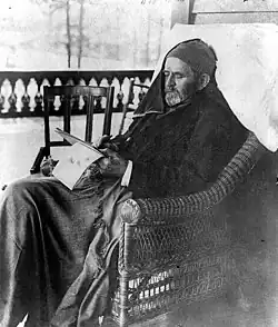 Photographie de Grant dans un fauteuil en osier sur le perron d'une maison écrivant sur un calepin. Il est en costume mais une couverture est posée sur ses jambes, il a un bonnet et un foulard cachant une partie de son visage.