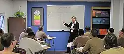 Une femme se tient devant une salle de classe et indique des fractions sur un tableau blanc.  Onze hommes sont assis à des bureaux en face d'elle.