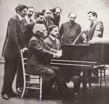 Photographie en noir et blanc d'un groupe d'hommes debout autour de deux hommes assis au piano.