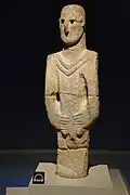 Statue en grès d'une huteur de 1,80 m6 dont les yeux sont représentés trous sculptés des obsidiennes noires. Un col ou bien un collier, a été taillé en relief sur le col de la statue. Les mains sont représentées serrées au niveau du corps de la statue, présentées sur le devant en couvrant les parties génitales.