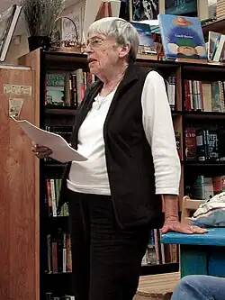 Photo en couleur de Le Guin en 2008 : c'est une femme âgée, aux cheveux blancs et courts, qui porte des lunettes, et se tient debout, en train de lire un texte écrit sur des feuilles de papier