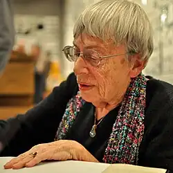 Gros plan d'une femme âgée avec des lunettes, des cheveux gris courts, et portant une écharpe écrit une dédicace sur un livre