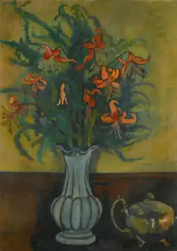 Un vase dans lequel on a mis des grandes plantes vertes ornées de fleurs rouges. À droite du vase, une théière métallique.