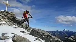 VTTiste descendant un chemin légèrement enneigé le long d'une montagne. À sa gauche, une croix a été installée au sommet de la montagne.