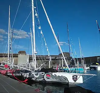 Vu de trois quarts avant, voilier blanc, gris et noir amarré au ponton. Un dragon à l'œil rouge est dessiné en grand sur la coque. Foils rouges.