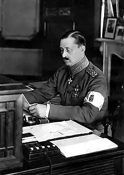 Photo en noir et blanc d'un homme moustachu lisant assis à une table, il porte un uniforme militaire et un brassard blanc au bras gauche