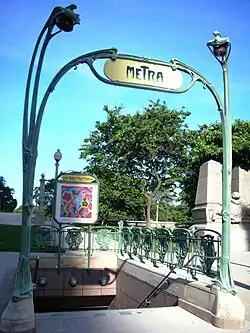 Photo couleur d'un entourage en fer forgé à écussons avec candélabres et écriteau marqué "metra" près d'un monument en pierre et sur fond de ciel bleu ciel et arbres au second plan